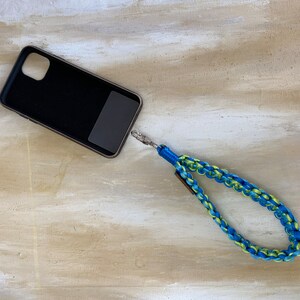 May include: Black phone case with a blue and yellow braided wrist strap. The strap is attached to the case with a silver-colored clasp. The phone case has a dark gray rectangle on the back.