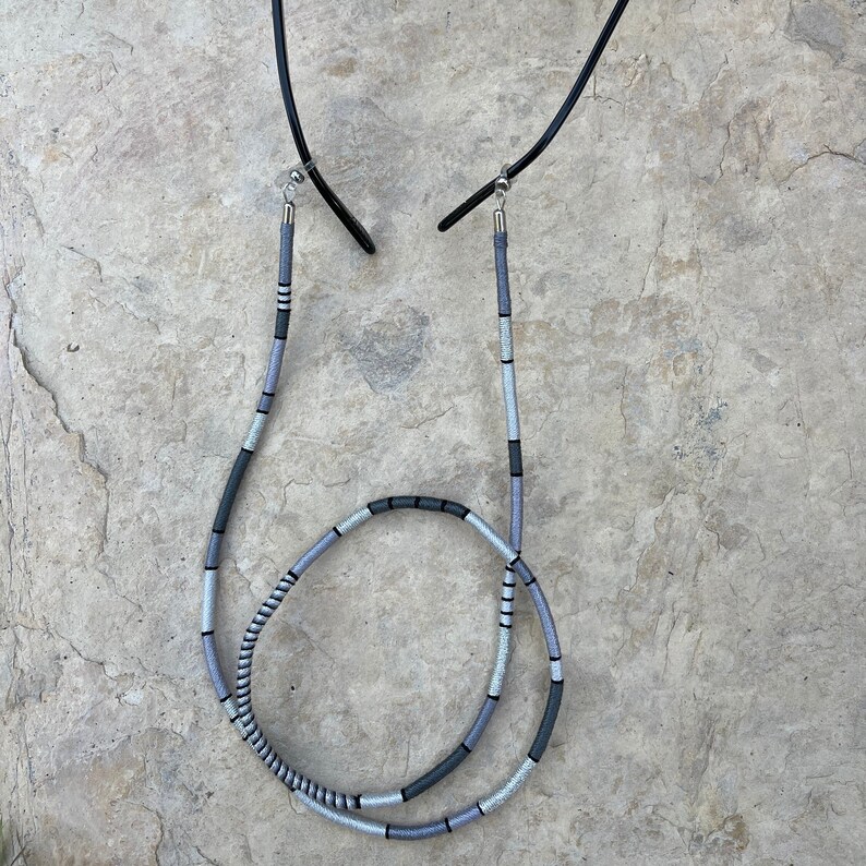 May include: A gray and black glasses chain with silver metal ends. The chain is made of woven cord and is designed to hold glasses around the neck.
