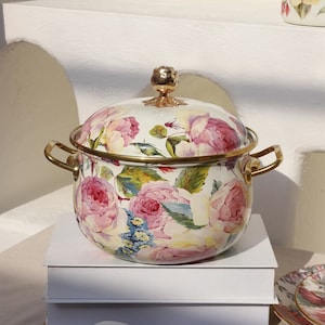 Op de afbeelding: Een witte emaille pot met een gouden deksel en handvatten. De pot is versierd met een bloemenpatroon van roze rozen en groene bladeren.