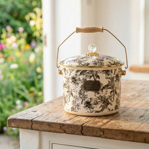Puede incluir: Bote de cocina color crema con un estampado floral en negro y gris. Tiene un borde y fijaciones de asa dorados, un asa de madera y un pomo de cristal transparente en la tapa. El bote está sobre una mesa de madera.