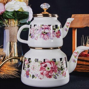 Puede incluir: Una tetera de esmalte blanco con un diseño floral y detalles en cuadros negros y blancos. La tetera tiene un borde dorado y un pomo de cristal en la tapa. La tetera está apilada sobre otra tetera a juego con un pico.