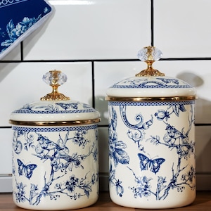 May include: Two white ceramic canisters with blue and gold floral designs and crystal knobs. The canisters are on a wooden surface.