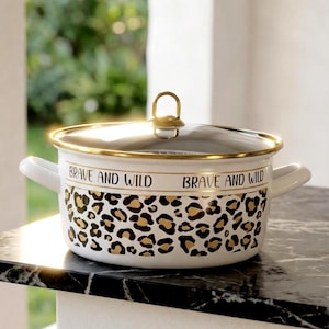 Op de afbeelding: Witte emaille kookpot met een gouden rand en handvat, met een luipaardprint ontwerp. De pot heeft de woorden "BRAVE AND WILD" erop gedrukt. De pot heeft twee witte handvatten en een doorzichtig deksel.