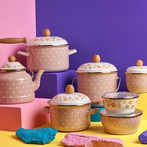 May include: A set of pink and white enamel cookware with a floral design and polka dots. The set includes a teapot, a large pot, two smaller pots, a bowl, and a lid. The cookware is arranged on a colourful background.