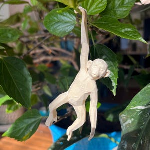Puede incluir: Una figura de mono de madera tallada en beige claro, colgando de una rama. El mono tiene los brazos y las piernas extendidos. El fondo presenta hojas verdes y una maceta azul, creando un entorno natural y decorativo.