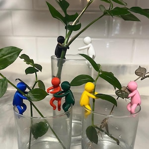 May include: A collection of clear glass vases holding green plants, each with a colorful figurine attached. The figurines are in various colors, including blue, red, green, yellow, pink, black, and white. The figurines appear to be holding the plant stems.