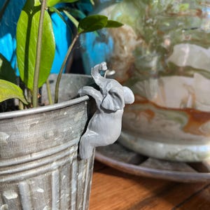 Puede incluir: Una figurita de elefante gris está unida al lado de una maceta de metal gris. La maceta está llena de una planta verde con hojas largas y delgadas.