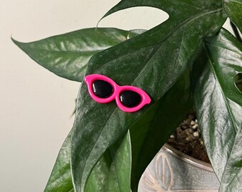 May include: Pink plastic sunglasses with black lenses on a green leaf.