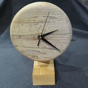Op de afbeelding: Een ronde houten klok met een natuurlijke lichtgekleurde houtnerf en donkere, organische spalted patronen over het hele oppervlak. De klok heeft drie dunne zwarte wijzers en staat op een rechthoekige houten voet voor verticale presentatie.