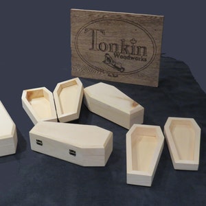 Peut inclure: Six petites boîtes en bois en forme de cercueil, toutes fermées, sur une surface gris foncé. Les boîtes sont en bois brut et sont toutes de la même taille. Une boîte en bois avec le texte "Tonkin Woodworks" est en arrière-plan.