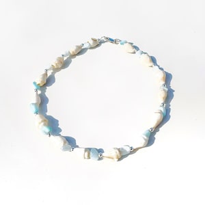Könnte beinhalten: Eine weiße und blaue Muschelkette mit silbernen Perlen. Die Kette ist aus natürlichen Materialien gefertigt und hat ein schlichtes, elegantes Design.
