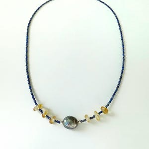 May include: A necklace made with small blue lapis lazuli beads and a single large grey pearl. The necklace also features small, flat, golden beads.