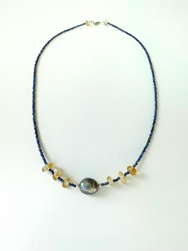 May include: A necklace made with small blue lapis lazuli beads and a single large grey pearl. The necklace also features small, flat, golden beads.