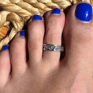 May include: A silver toe ring with a wave design is worn on a foot with blue nail polish. The foot rests on a woven, tan surface. The ring is detailed with a repeating pattern of waves.