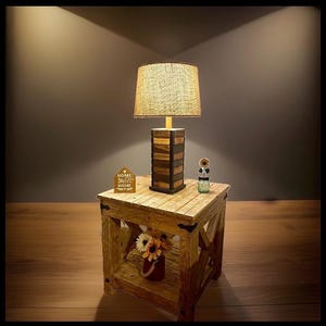 May include: A wooden side table with an X-frame design and a lower shelf holding a small flower arrangement. On top sits a lamp with a textured beige shade and a stacked wooden base, a small house-shaped sign reading Home Sweet Home, and a sunflower.