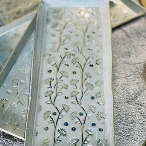 Puede incluir: Un juego de tres bandejas rectangulares de cerámica con un esmalte azul claro y un diseño floral en relieve. El diseño presenta delicadas flores blancas y puntos azules.