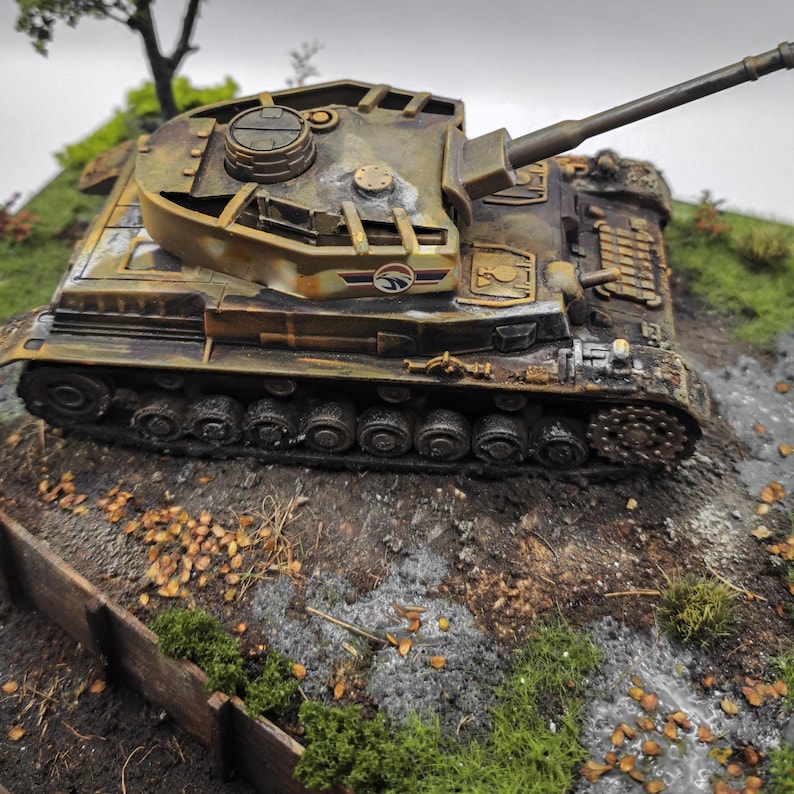 May include: A detailed model of a military tank, painted in shades of brown, green, and yellow, with a long gun barrel. The tank is positioned on a miniature terrain with grass, leaves, and a small wooden fence, creating a realistic diorama.
