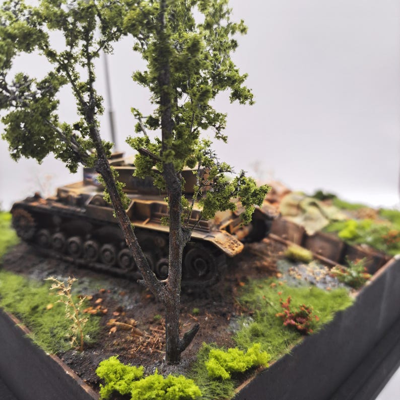 May include: A detailed model of a military tank, painted in shades of brown and tan, sits amidst a miniature landscape. The scene includes a small tree with green foliage, patches of grass, and other miniature details, creating a realistic diorama.