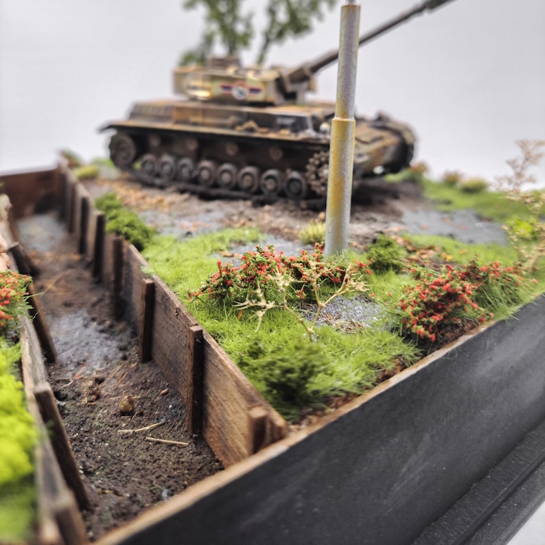 May include: A detailed diorama of a military scene featuring a model tank, a trench, and vegetation. The tank is painted in camouflage colors. The trench is made of wooden planks. Green grass and red-flowered bushes add color.