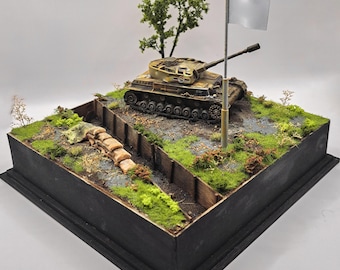 May include: A detailed diorama of a military scene featuring a tan tank on a grassy hill. A trench with sandbags and a small tree are in the foreground. A white flag flies from a pole, suggesting surrender. The diorama is set in a black wooden box.