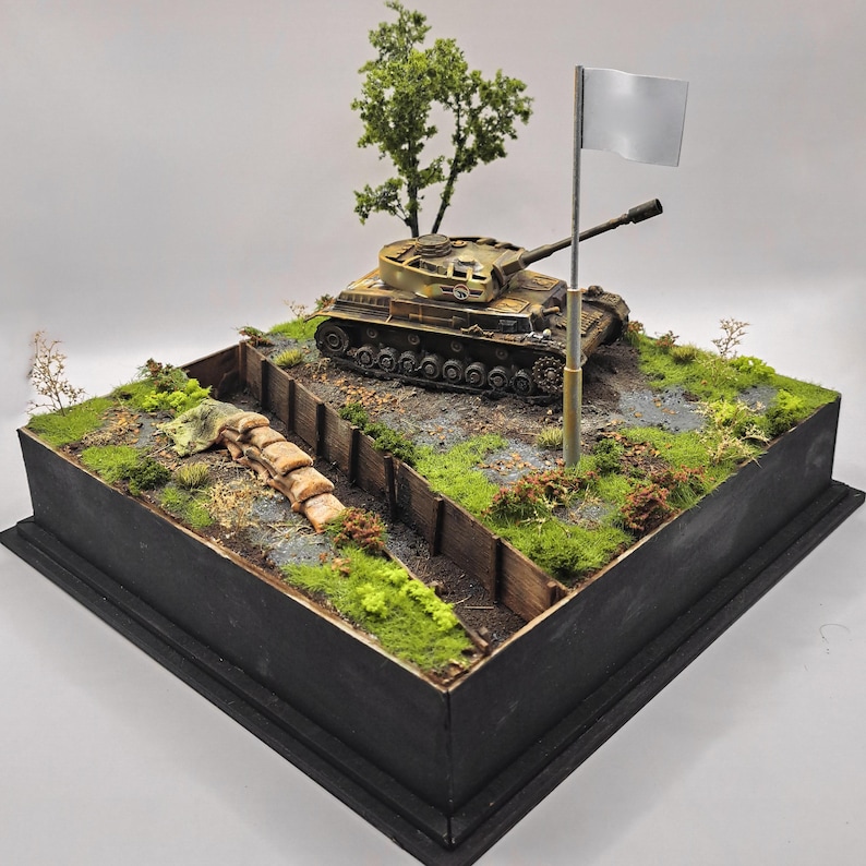 May include: A detailed diorama of a military scene featuring a tan tank on a grassy hill. A trench with sandbags and a small tree are in the foreground. A white flag flies from a pole, suggesting surrender. The diorama is set in a black wooden box.