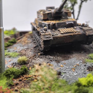 May include: A detailed miniature model of a military tank, showcasing a weathered, battle-worn appearance. The tank is painted in shades of brown and green, with visible rust and dirt. The model sits on a diorama base with grass and muddy terrain.