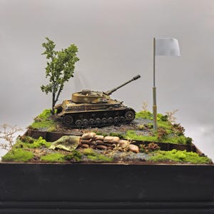 May include: A detailed model of a military tank in a diorama setting. The tank is painted in camouflage colors and sits on a terrain of grass, dirt, and small plants. A white flag is raised nearby, suggesting surrender.