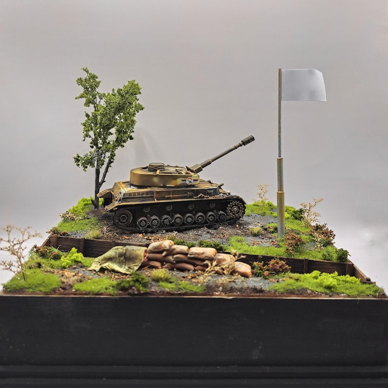 May include: A detailed model of a military tank in a diorama setting. The tank is painted in camouflage colors and sits on a terrain of grass, dirt, and small plants. A white flag is raised nearby, suggesting surrender.