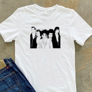 May include: White t-shirt with a black and white silhouette of a band. The band members are all wearing different clothing and have different hairstyles. The t-shirt is a great way to show your love for the band.