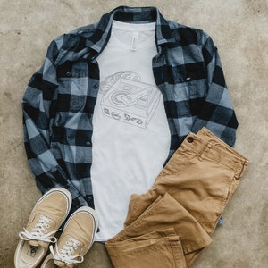 Op de afbeelding: Een casual outfit met een wit t-shirt met een platenspeler-afbeelding, een blauw-zwart geruit flanellen overhemd, een kaki broek en beige sneakers met witte veters. De items zijn gerangschikt op een neutrale ondergrond.