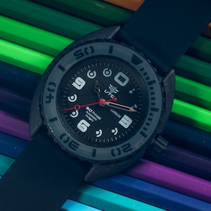 Op de afbeelding: Een zwart polshorloge met een grijze bezel en een zwarte band. De wijzerplaat van het horloge is zwart met witte cijfers en een rode secondewijzer. De wijzerplaat van het horloge heeft ook de tekst "3D Printed Automatic Hungary" en "CARBON".