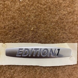 Op de afbeelding: Een zilverkleurig "EDITION 1" embleem op een witte achtergrond. Het embleem heeft een metallic afwerking en is geplaatst tegen een bruine, getextureerde achtergrond. De tekst is vetgedrukt, sans-serif.