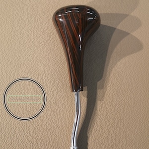 Puede incluir: Una perilla de cambio de marchas de madera con un patrón de rayas marrones oscuras y negras. La perilla está unida a una palanca de cambio de marchas de metal cromado.