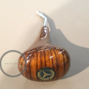 Op de afbeelding: Een houten versnellingspookknop met een Mercedes-Benz-logo. De knop is bruin met een houtnerfpatroon en heeft een chromen hendel.