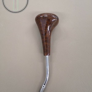 Könnte beinhalten: Ein brauner Holz- und Chrom-Schalthebelknauf. Der Knauf ist rund und hat eine Holzmaserung. Der Chromschaft ist gebogen.