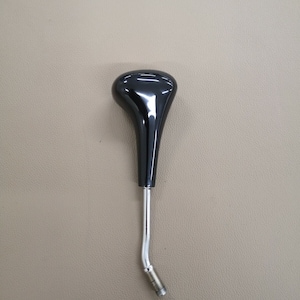 Könnte beinhalten: Schwarzer Schaltknauf mit einem verchromten Metallfuß. Der Knauf ist tropfenförmig und hat eine glatte, glänzende Oberfläche.