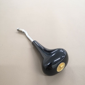 Pode incluir: Um botão de mudança de marcha preto com um detalhe dourado e um braço de metal prateado. O botão tem um acabamento brilhante e uma forma arredondada. O detalhe dourado é um pequeno design geométrico.