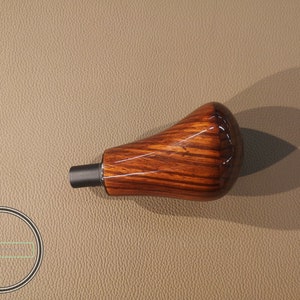 以下が含まれることがあります： 光沢仕上げの木製ギアシフトノブ。ノブは温かみのある茶色で、木目がはっきりと見え、ベースは黒です。自動車用途向けに設計された製品です。