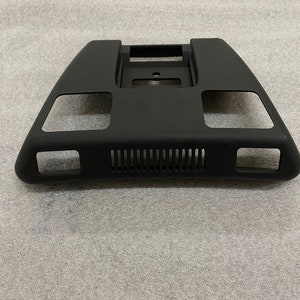 May include: Black plastic car part with a rectangular opening and a series of small holes. The part has a curved shape and is designed to fit into a car's interior.