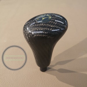 Puede incluir: Una palanca de cambios de fibra de carbono negra con un logotipo dorado de Mercedes-Benz.