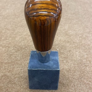 Puede incluir: Una palanca de cambios de madera con una base de metal plateado y un logotipo de Mercedes-Benz en la parte superior. La palanca está sobre un cuadrado de terciopelo azul.