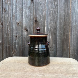 May include: A handcrafted ceramic jar with a lid. The jar is dark brown with a glossy finish and two small handles. The lid is a lighter brown with a small knob on top. The jar sits on a light-colored wooden surface.
