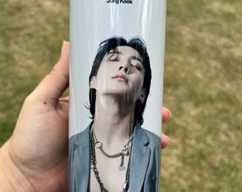 May include: White tumbler with a photo of a man with black hair and a gray jacket. The text "SEVEN Jung Kook" is printed on the tumbler.