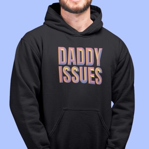 Könnte beinhalten: Schwarzer Kapuzenpullover mit dem Text "DADDY ISSUES" in einer bunten, 3D-Schrift.