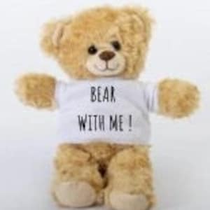 May include: A plush, light brown teddy bear wearing a white t-shirt. The t-shirt has the text "BEAR WITH ME!" in black. The bear has a stitched black nose and dark eyes. The bear's arms are outstretched.