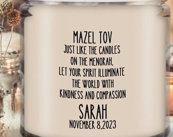 May include: A glass jar candle with a black lid. The candle has a white label with the text "MAZEL TOV" and a personalized message. The candle is surrounded by decorative elements, including dried flowers and a small lit candle.