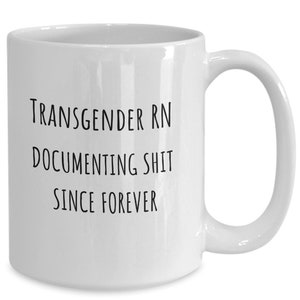 Könnte beinhalten: Weißer Keramikbecher mit gebogenem Henkel. Der Becher hat einen schwarzen Text mit der Aufschrift "TRANSGENDER RN DOCUMENTING SHIT SINCE FOREVER". Der Becher ist für heiße oder kalte Getränke geeignet.