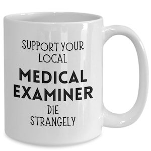 Op de afbeelding: Witte keramische mok met de zwarte tekst "SUPPORT YOUR LOCAL MEDICAL EXAMINER DIE STRANGELY". De mok heeft een gebogen handvat en is ontworpen voor warme dranken.