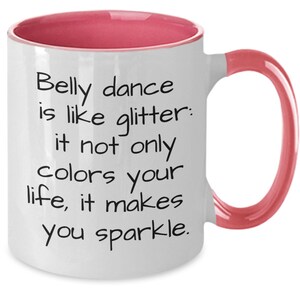 May include: White ceramic mug with a pink interior and handle. The mug features the text "Belly dance is like glitter: it not only colors your life, it makes you sparkle" in black lettering.