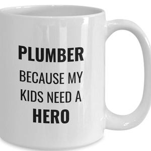 May include: White ceramic mug with the text "PLUMBER BECAUSE MY KIDS NEED A HERO" in black, bold letters. The mug has a curved handle and a smooth, glossy finish. A practical and humorous gift.